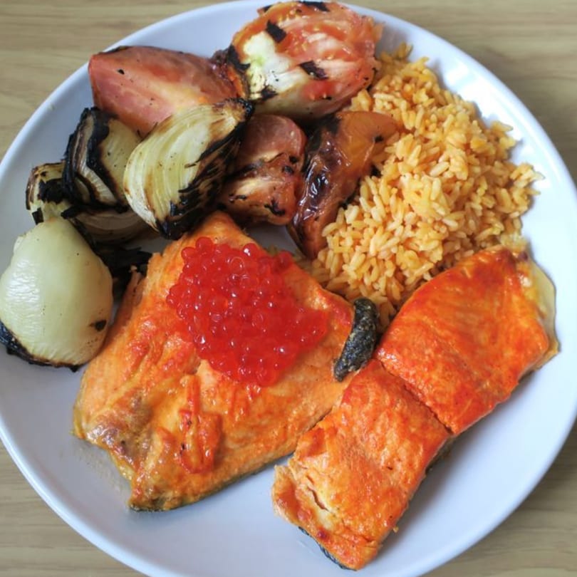 Grilled salmon with red caviar, roasted onions, tomatoes, and rice on a white plate.